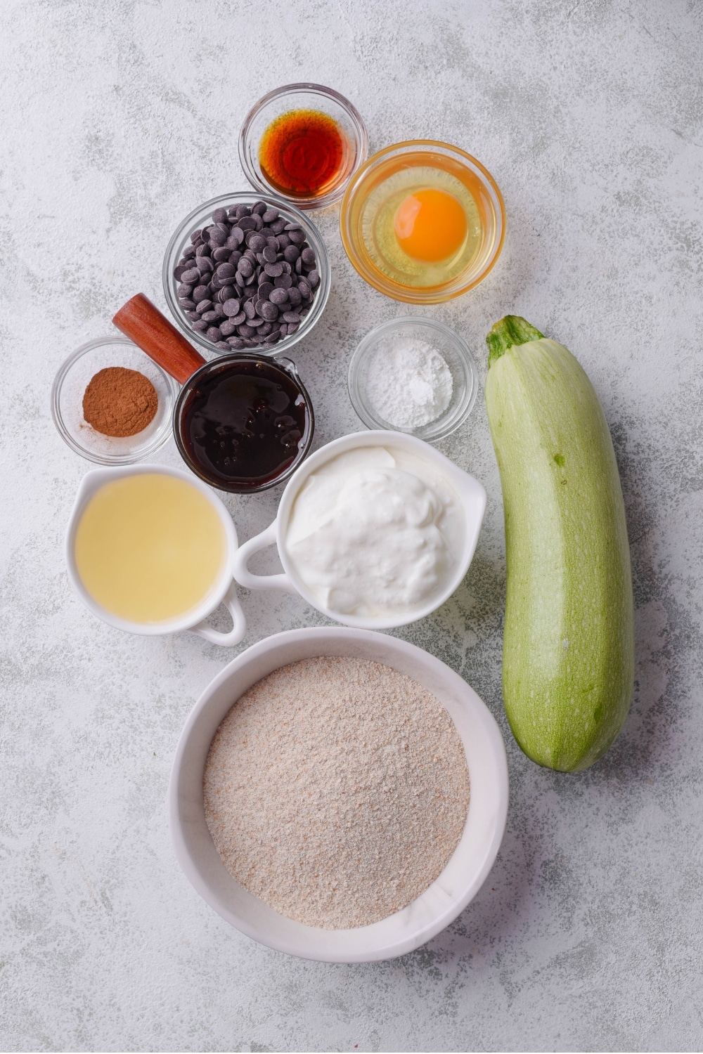 An overhead shot of multiple bowls carrying a variety of ingredients including egg, yogurt, chocolate chips, and flour. A zucchini is next to the bowls.