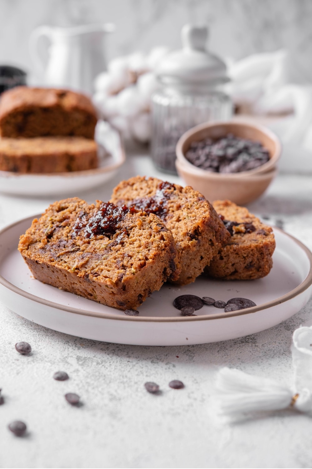 Three slices of low calorie zucchini bread with chocolate chips arranged on a white ceramic plate. There are a few chocolate chips scattered with another loaf of bread and bowl of chocolate chips in the background.