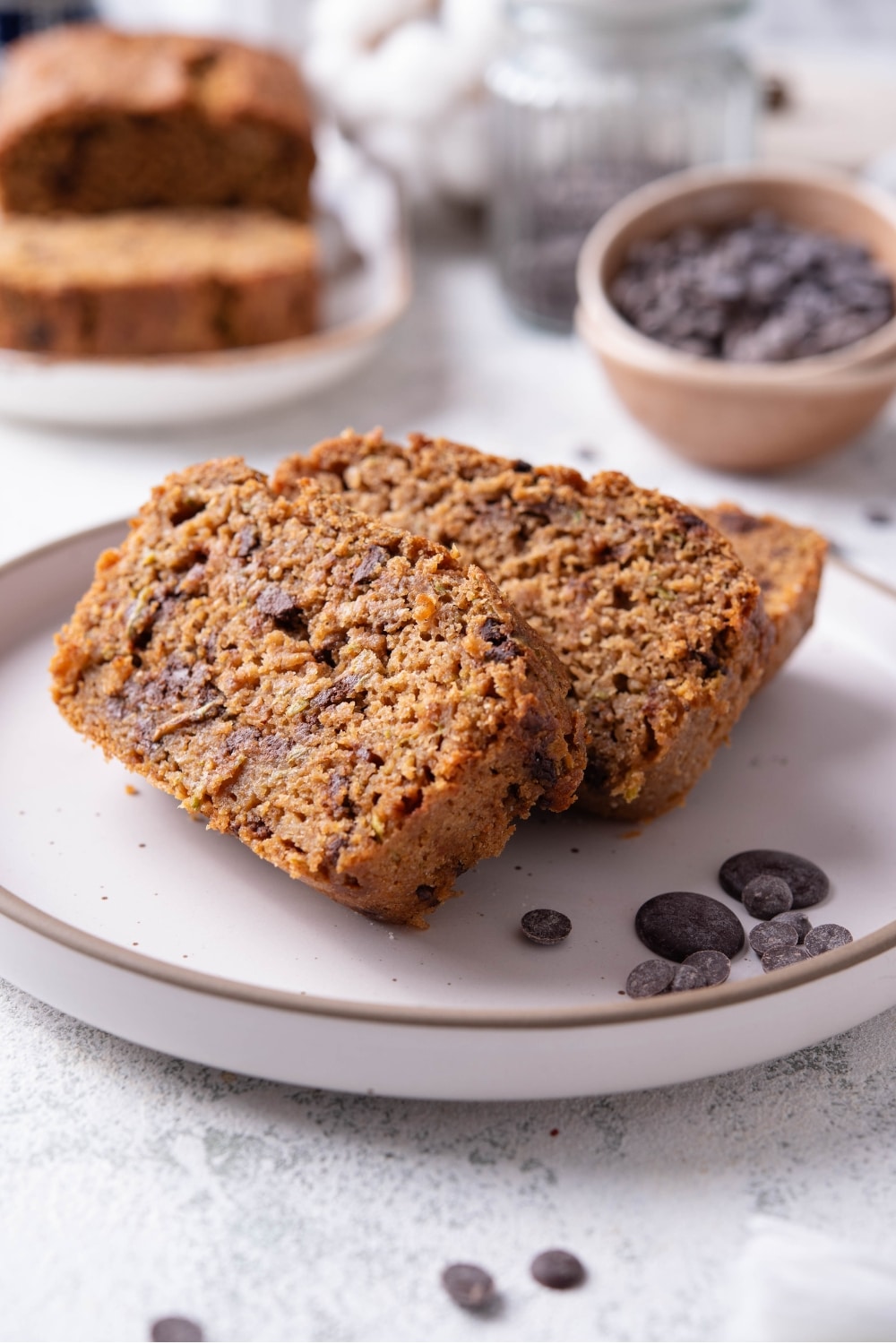 Three slices of low calorie zucchini bread with chocolate chips arranged on a white ceramic plate. There are a few chocolate chips scattered with another loaf of bread and bowl of chocolate chips in the background.