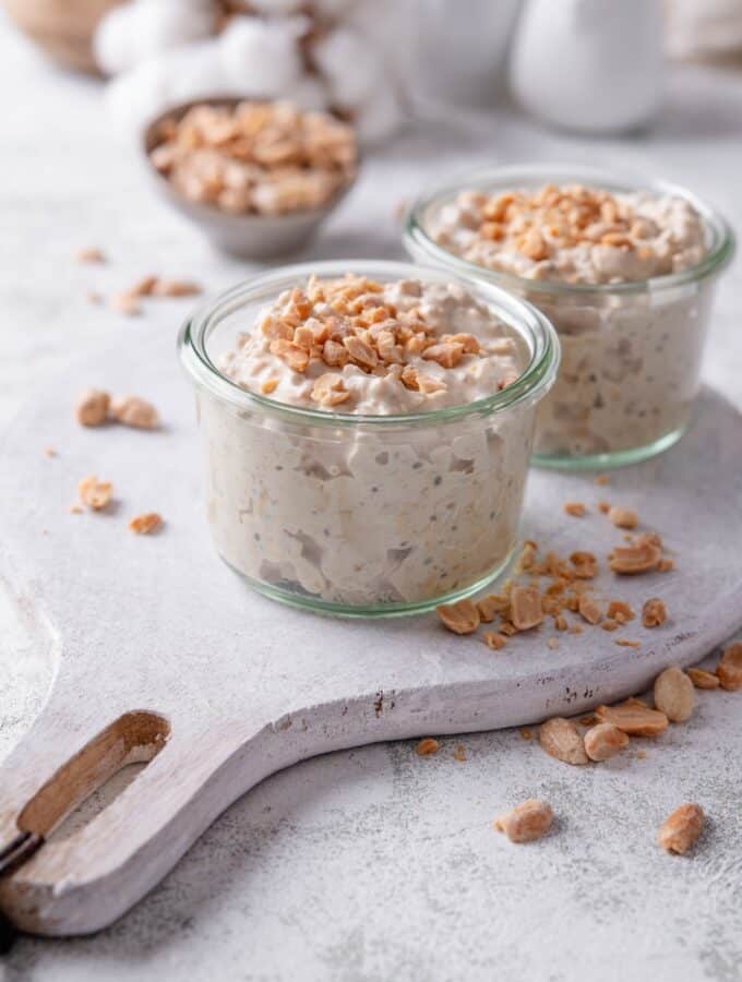 Two small glass jars filled with peanut butter overnight oats on top of a wooden serving board with peanuts sprinkled around it. There are peanuts sprinkled on top of each jar, and additional peanuts sprinkled around the jars.