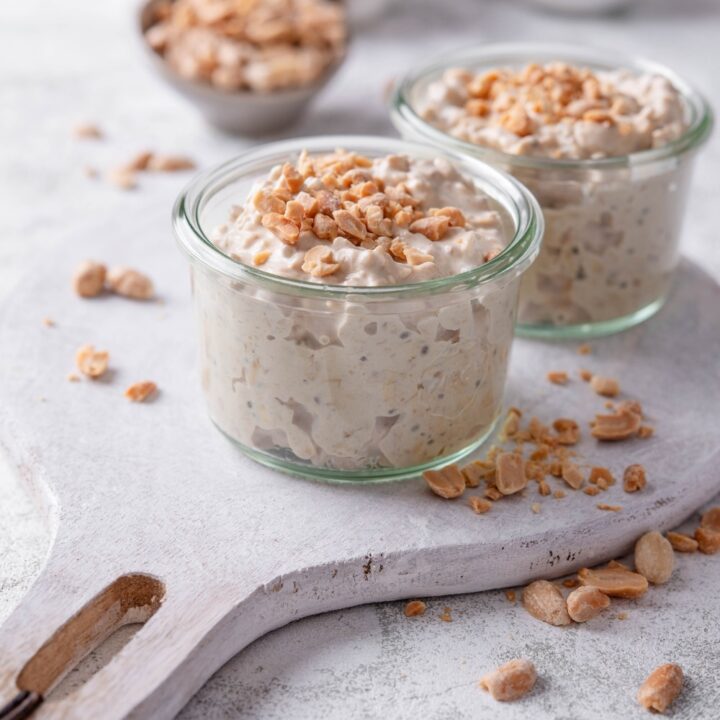 Two small glass jars filled with peanut butter overnight oats on top of a wooden serving board with peanuts sprinkled around it. There are peanuts sprinkled on top of each jar, and additional peanuts sprinkled around the jars.