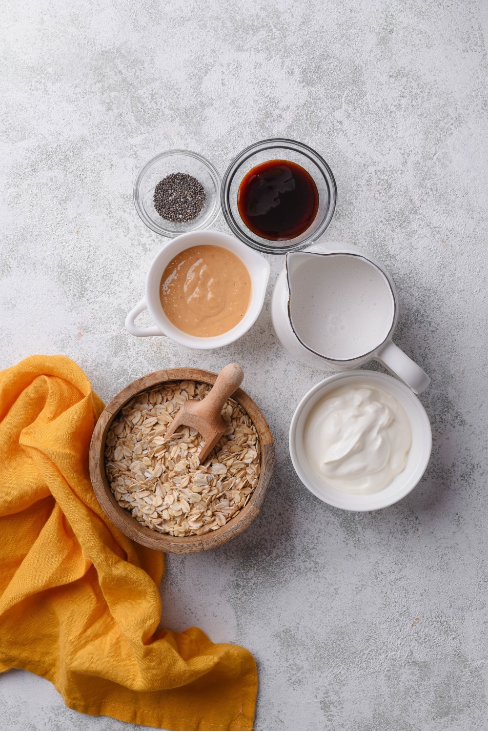 An overhead shot of several bowls in various shapes and sizes filled with ingredients for peanut butter overnight oats including almond milk, greek yogurt, chocolate sauce, chia seeds, peanut butter, and oats.