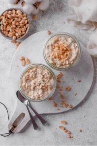 An overhead shot of two glass jars filled with peanut butter overnight oats. One jar has peanuts crumbled on top, with a bowl of peanuts to the side. The jars are on top of a serving board with two spoons on the side.