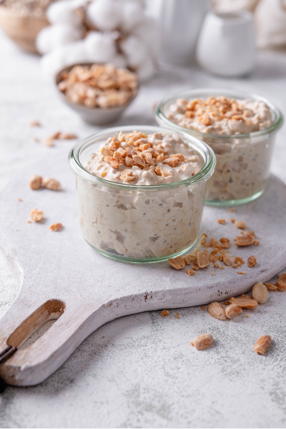 Two small glass jars filled with peanut butter overnight oats on top of a wooden serving board with peanuts sprinkled around it. There are peanuts sprinkled on top of each jar, and additional peanuts sprinkled around the jars.