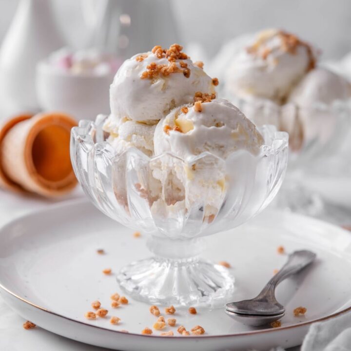 Three scoops of vanilla protein ice cream in a glass sundae bowl on a white plate with a spoon to the side. There are some nuts sprinkled on top.