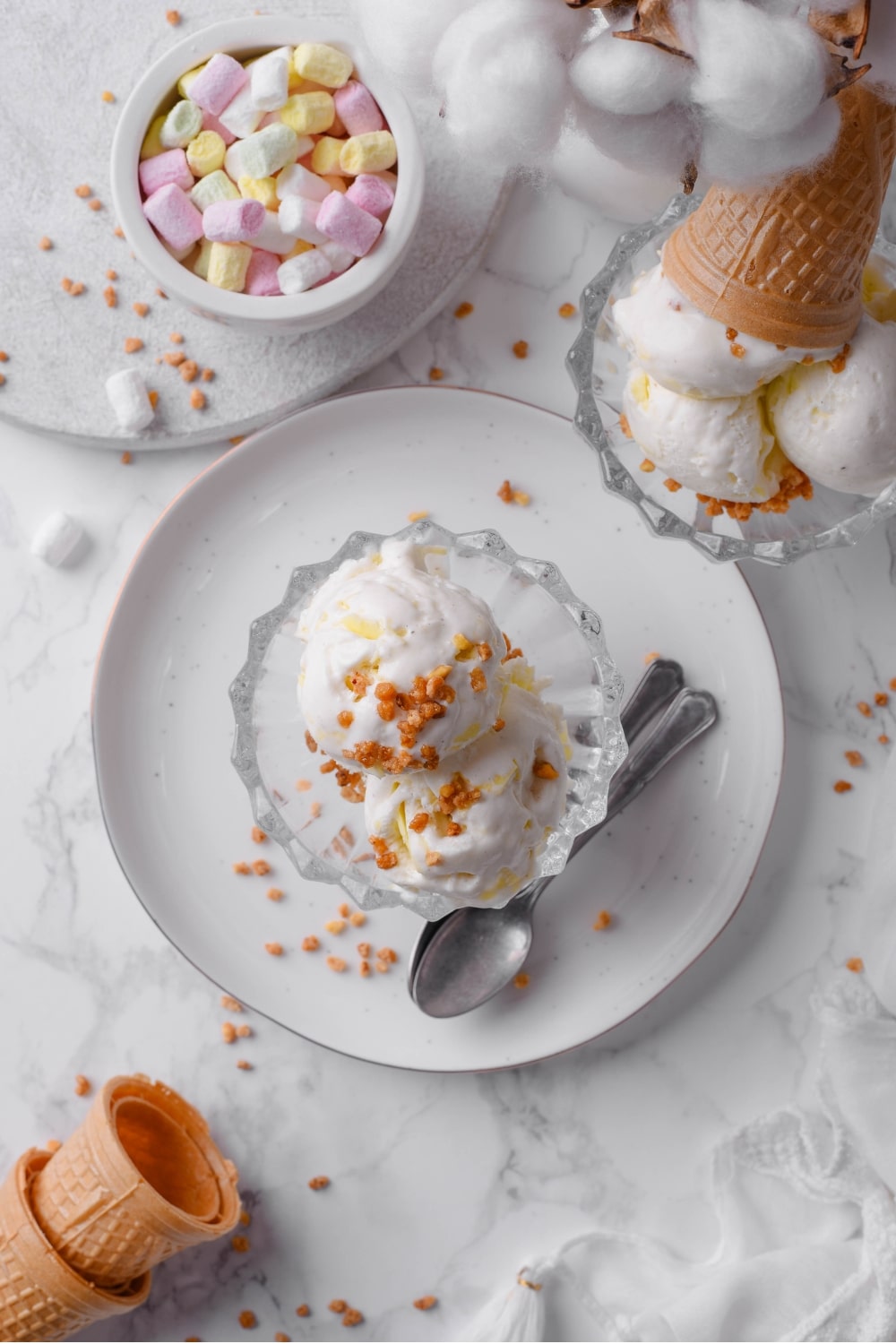 An overhead shot of vanilla protein ice cream in a glass sundae bowl on a white plate. There is a second sundae bowl with ice cream on the side, as well as some ice cream cones and marshmallows surrounding the plate.