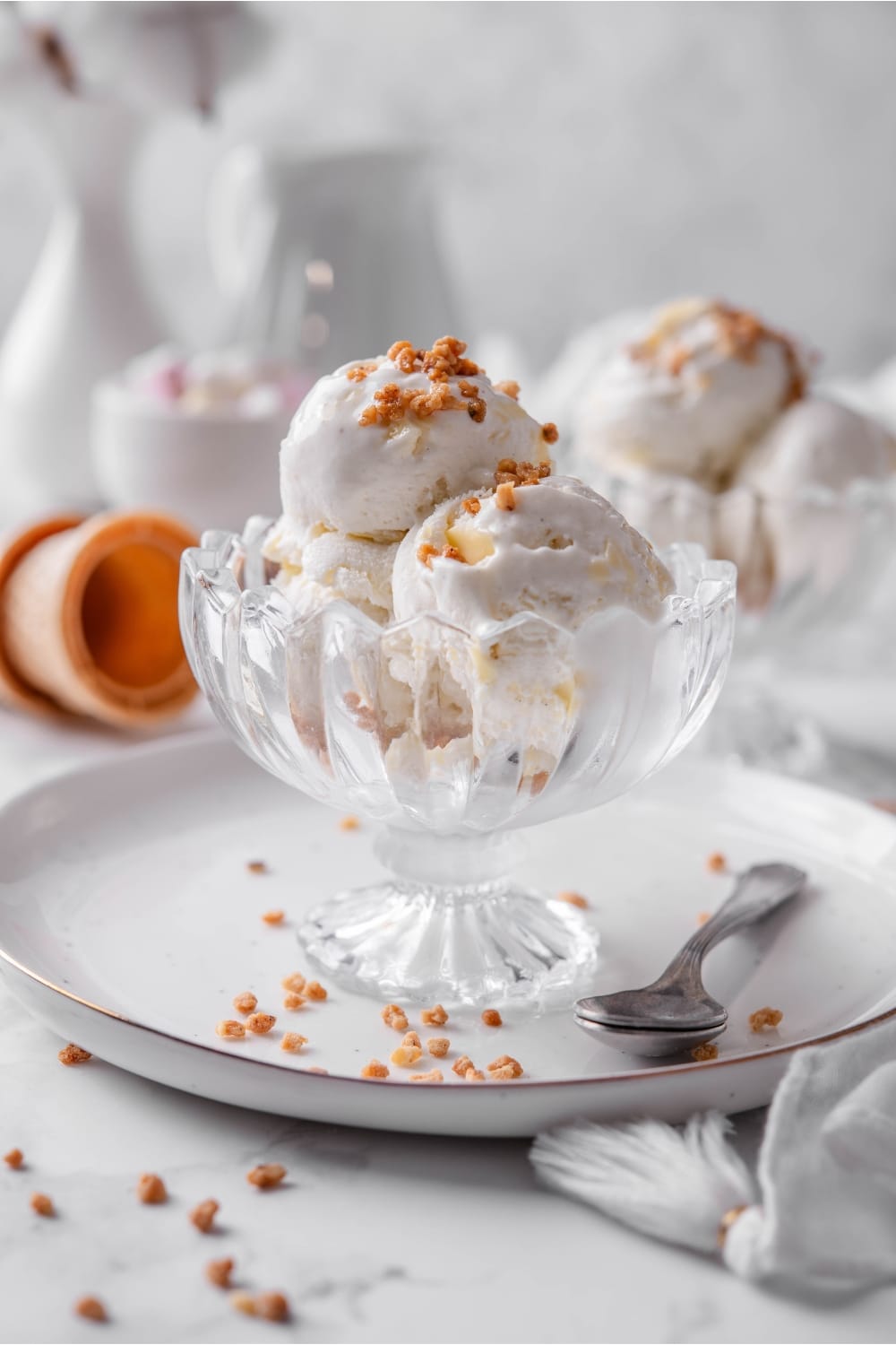 Three scoops of vanilla protein ice cream in a glass sundae bowl on a white plate with a spoon to the side. There are some nuts sprinkled on top.