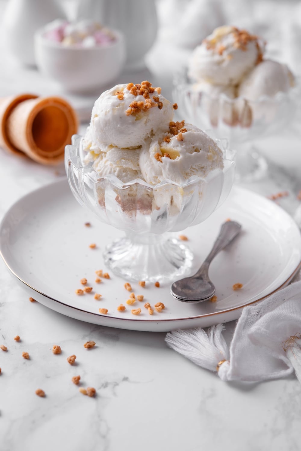 Three scoops of vanilla protein ice cream in a glass sundae bowl on a white plate with a spoon to the side. There are some nuts sprinkled on top.