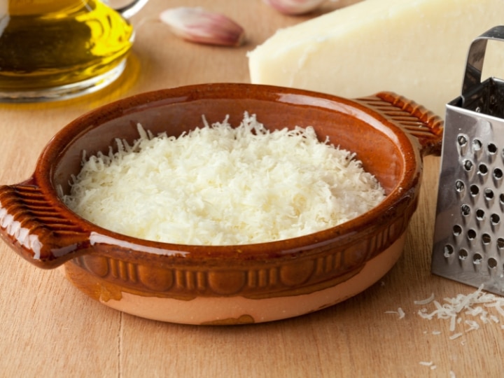 A bowl of romano cheese on a wood table with a cheese grater next to it.