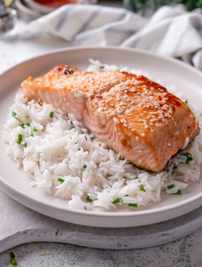 A white ceramic plate with teriyaki salmon served on top of white rice with sesame seeds and chopped chives. The plate rests on a wooden serving board with a striped napkin in the background