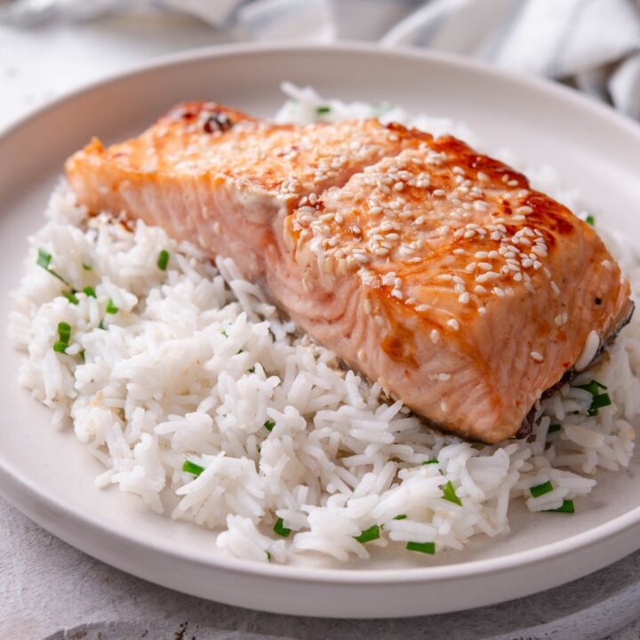 A white ceramic plate with teriyaki salmon served on top of white rice with sesame seeds and chopped chives. The plate rests on a wooden serving board with a striped napkin in the background