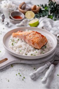 A white ceramic plate with teriyaki salmon served on top of white rice with sesame seeds and chopped chives. The plate rests on a wooden serving board with a fork and knife next to it.