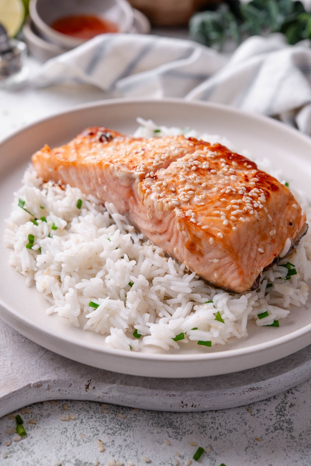 A white ceramic plate with teriyaki salmon served on top of white rice with sesame seeds and chopped chives. The plate rests on a wooden serving board with a striped napkin in the background