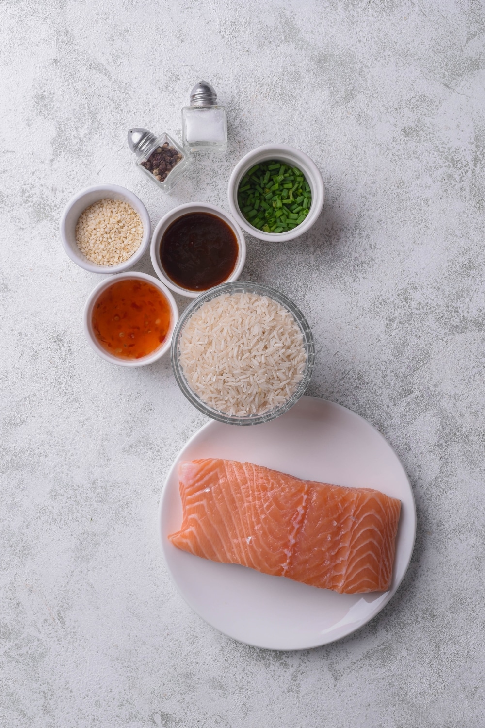 An overhead shot of various bowls of ingredients for salmon and rice including salmon, rice, teriyaki sauce, chili sauce, sesame seeds, and chives