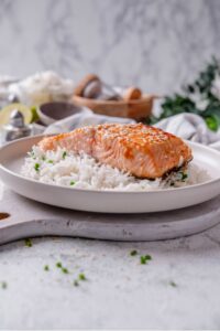A white ceramic plate with teriyaki salmon and sesame seeds served on top of white rice with chopped chives. The plate rests on a wooden serving board with wooden bowls in the background.