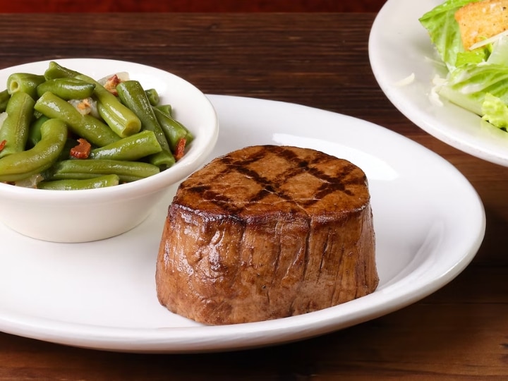 A piece of streak on a white plate with a bowl of green beans next to it.