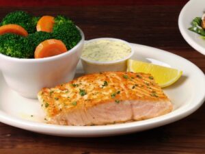 A piece of salmon on a white plate with a white bowl of broccoli and carrots.