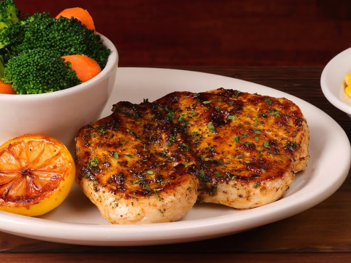 A piece of herb crusted chicken on a white plate with a bowl of broccoli and carrots on it.