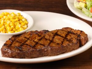 A piece of New York strip steak on a white plate with a bowl of corn on it too.
