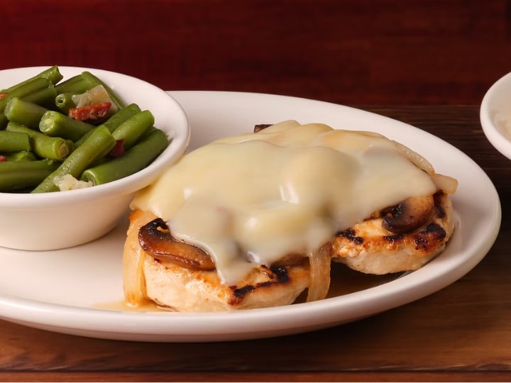 Melted cheese on top of a grilled chicken breast on a white plate with a bowl of green beans.