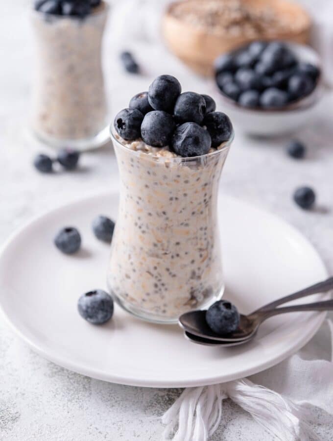 A glass jar filled with overnight oats and topped with fresh blueberries. The jar is on top of a ceramic plate with blueberries scattered around it, and a spoon in front.