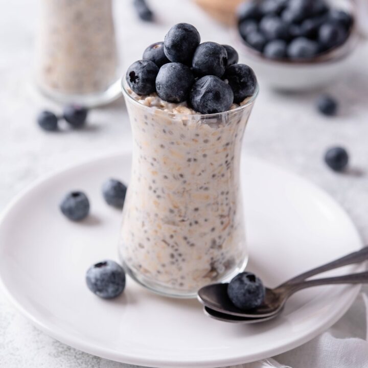 A glass jar filled with overnight oats and topped with fresh blueberries. The jar is on top of a ceramic plate with blueberries scattered around it, and a spoon in front.