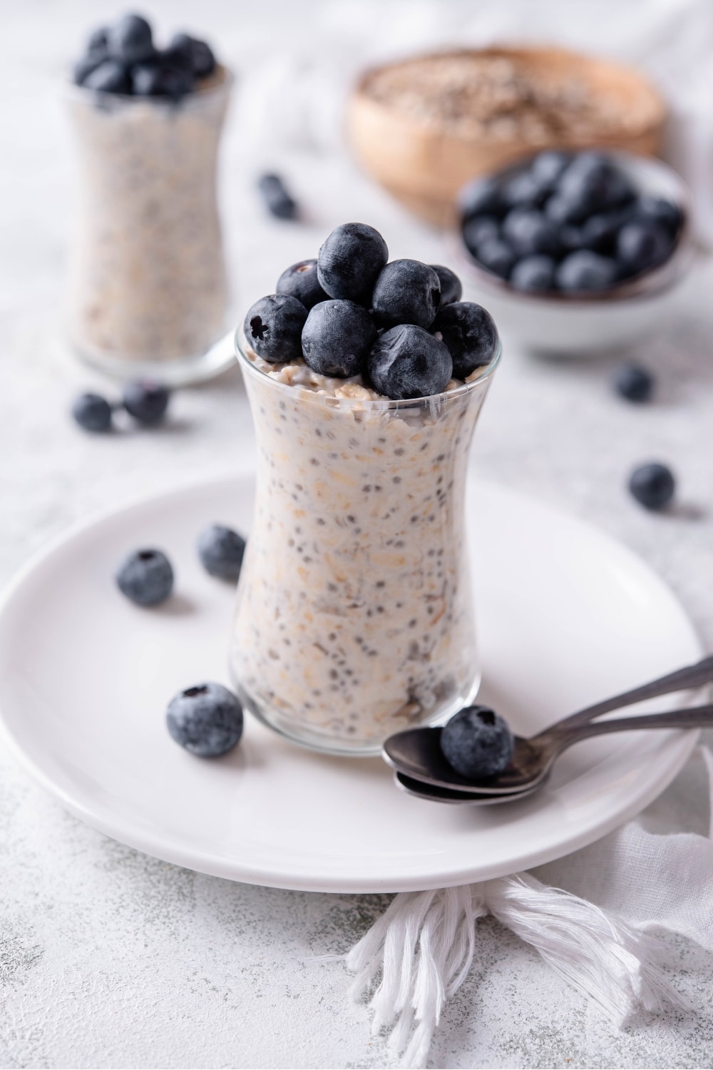 A glass jar filled with overnight oats and topped with fresh blueberries. The jar is on top of a ceramic plate with blueberries scattered around it, and a spoon in front.