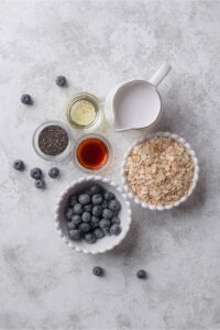 Several bowls of various sizes containing overnight oats ingredients such as blueberries, oats, chia seeds, almond milk, and vanilla extract.