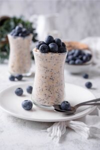A glass jar filled with overnight oats and topped with fresh blueberries. The jar is on top of a ceramic plate with blueberries scattered around it, and a spoon in front.