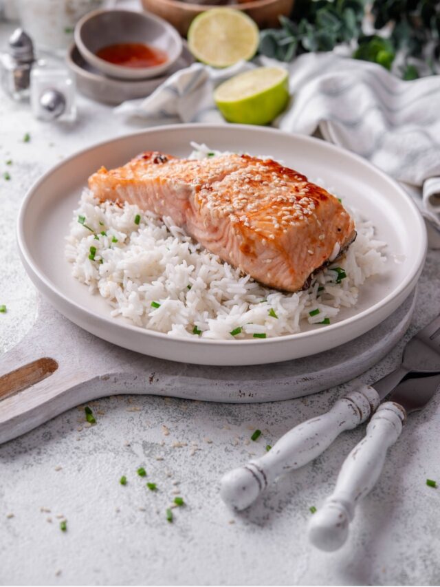 A white ceramic plate with teriyaki salmon served on top of white rice with sesame seeds and chopped chives. The plate rests on a wooden serving board with a fork and knife next to it.