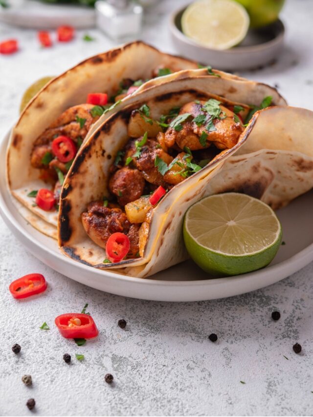A plate with two chicken al pastor tacos with a halved lime on the side. The tacos contain chicken, pineapple, and peppers with cilantro on top. There is a bowl limes in the background.