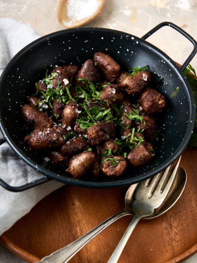 A ceramic bowl filled with cooked chicken hearts topped with fresh parsley. The bowl is on a wooden server with a fork, spoon, and napkin next to the bowl.