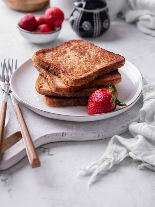 Four slices of french toast stacked on a white plate on top of a serving board. There is a strawberry and forks on the side.