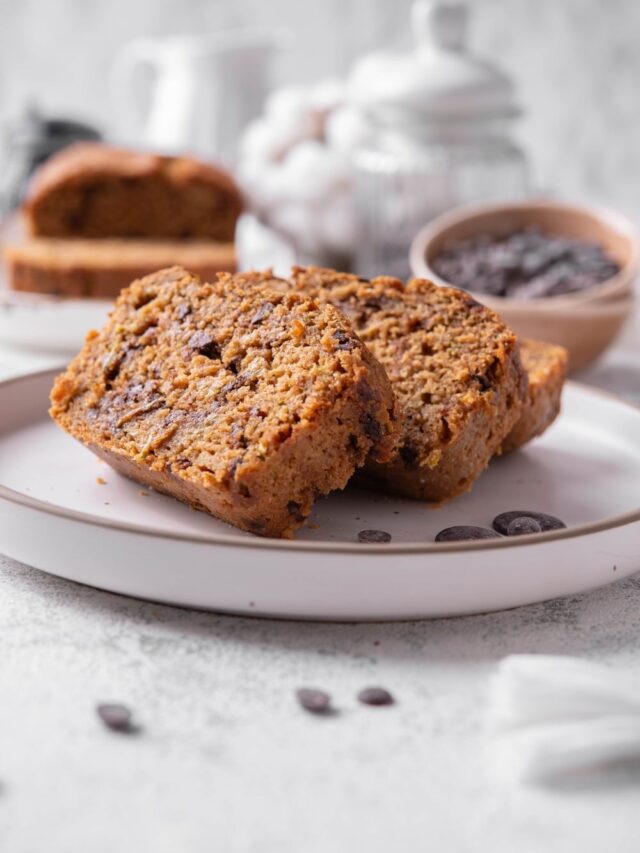 Three slices of low calorie zucchini bread with chocolate chips arranged on a white ceramic plate. There are a few chocolate chips scattered with another loaf of bread and bowl of chocolate chips in the background.