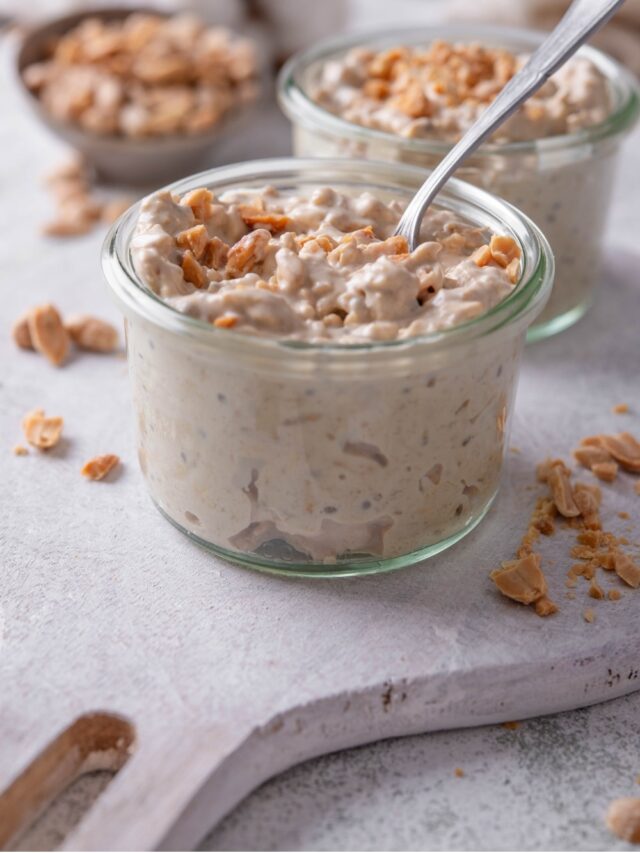 A small glass jar filled with peanut butter overnight oats on top of a wooden serving board with peanuts sprinkled around it. There is a spoon dipped into the jar.