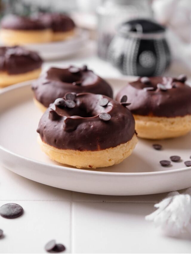 A ceramic plate with three baked donuts with chocolate frosting on top. The donuts are sprinkled with chocolate chips and there is an additional plate of donuts in the background.