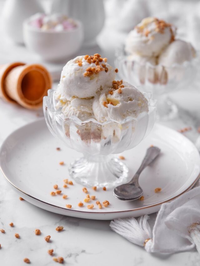 Three scoops of vanilla protein ice cream in a glass sundae bowl on a white plate with a spoon to the side. There are some nuts sprinkled on top.