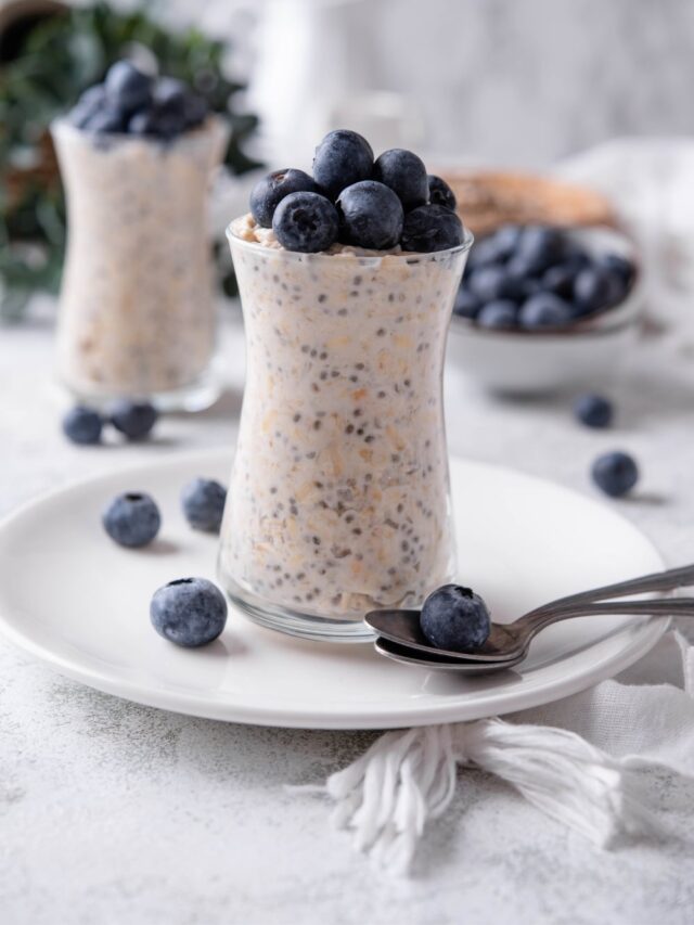 A glass jar filled with overnight oats and topped with fresh blueberries. The jar is on top of a ceramic plate with blueberries scattered around it, and a spoon in front.