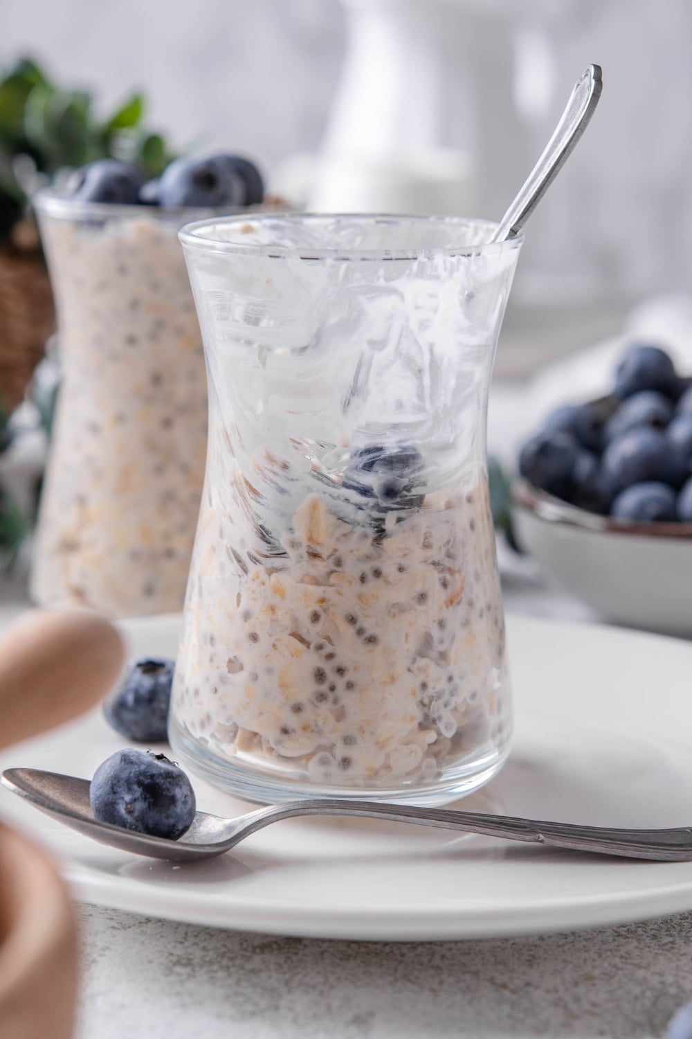 A half- eaten glass jar filled with overnight oats and topped with fresh blueberries. The jar is on top of a ceramic plate with blueberries scattered around it, and a spoon in front.