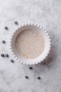 An overhead shot of a ceramic bowl filled with an oat, chia seed, and milk mixture on a marble counter with a few scattered blueberries surrounding it.