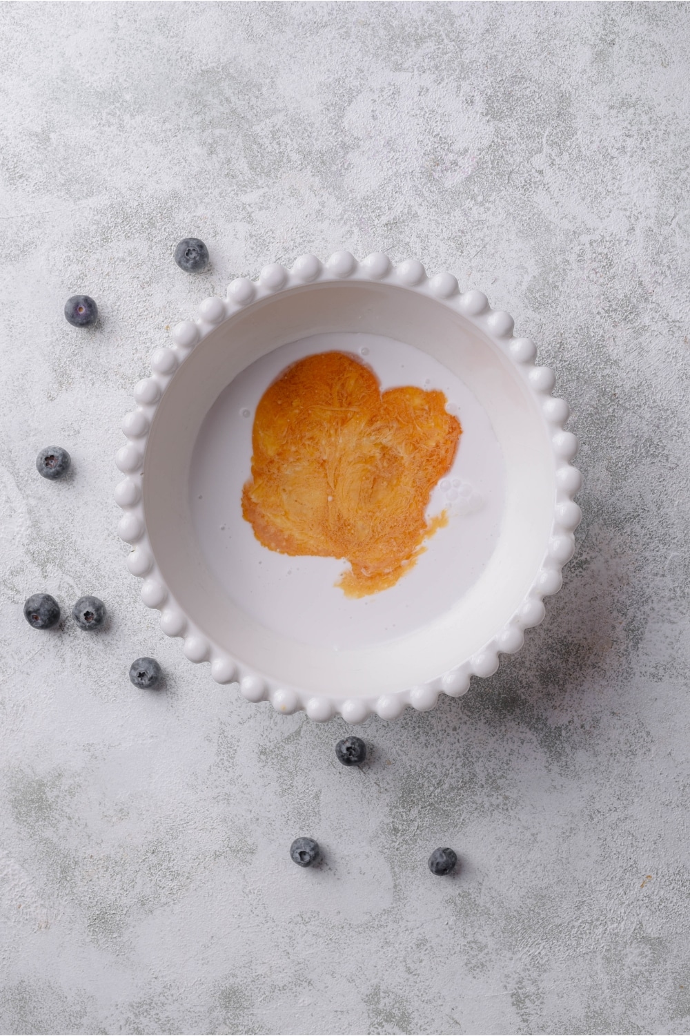 A ceramic bowl filled with milk, vanilla, and almond extract. The bowl rests on a marble counter with some blueberries scattered around it.