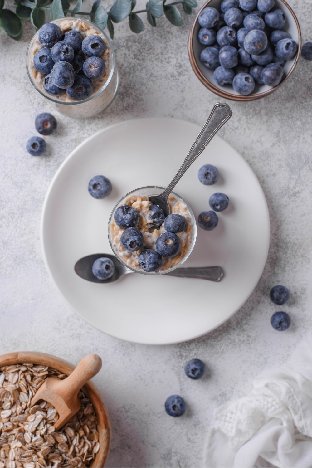 An overhead shot of a glass jar filled with overnight oats and fresh blueberries on top. The jar is on top of a ceramic plate with blueberries surrounding it and a spoon inserted in the oats.