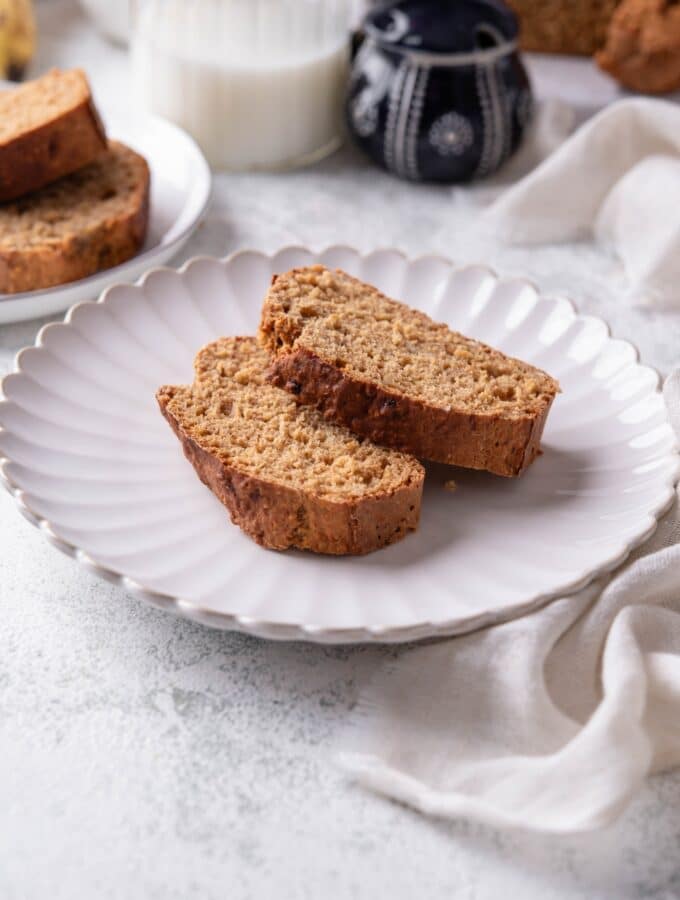 Two slices of low calorie banana bread on a white ceramic plate. There is a cloth napkin underneath is and an additional plate of banana bread slices in the background.