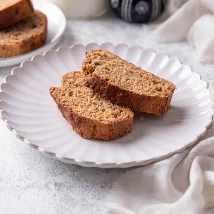 Two slices of low calorie banana bread on a white ceramic plate. There is a cloth napkin underneath is and an additional plate of banana bread slices in the background.