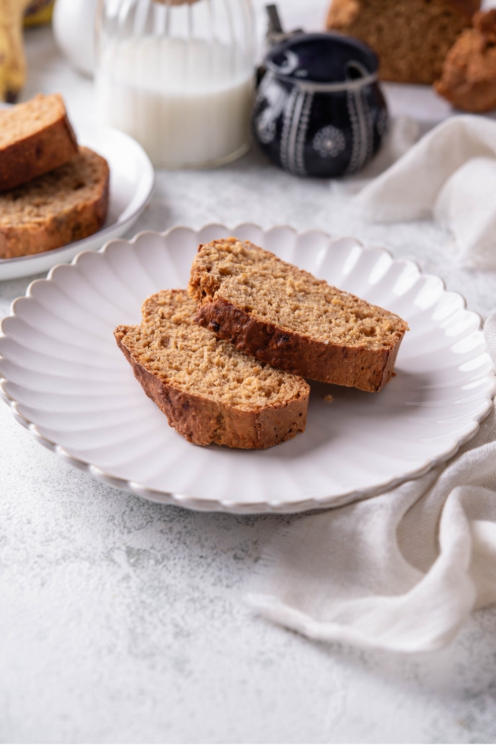 Two slices of low calorie banana bread on a white ceramic plate. There is a cloth napkin underneath is and an additional plate of banana bread slices in the background.