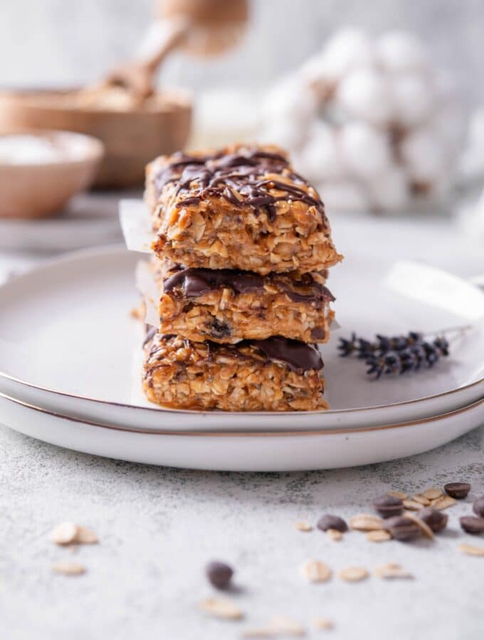 Three low calorie energy bars with chocolate drizzle stacked on a white ceramic plate. There are chocolate chips and oats sprinkled in front of the plate.