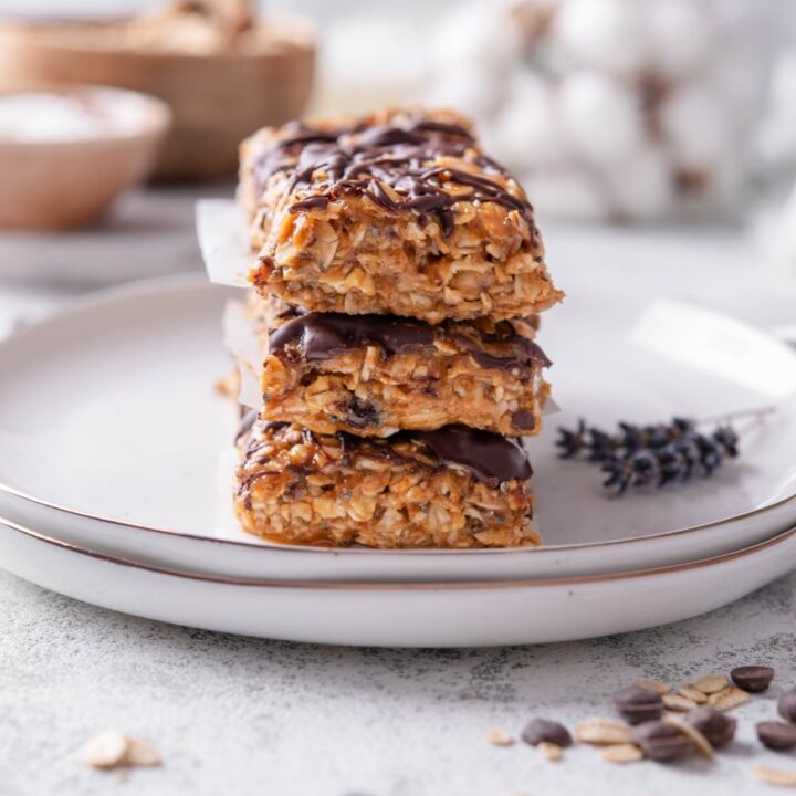 Three low calorie energy bars with chocolate drizzle stacked on a white ceramic plate. There are chocolate chips and oats sprinkled in front of the plate.