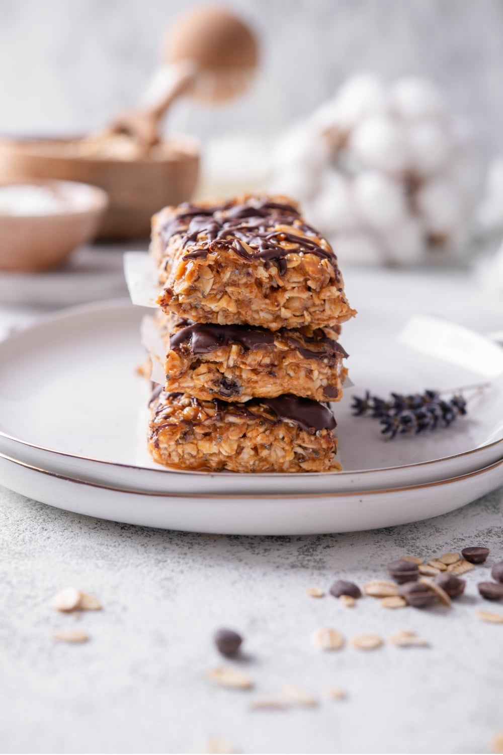 Three low calorie energy bars with chocolate drizzle stacked on a white ceramic plate. There are chocolate chips and oats sprinkled in front of the plate.