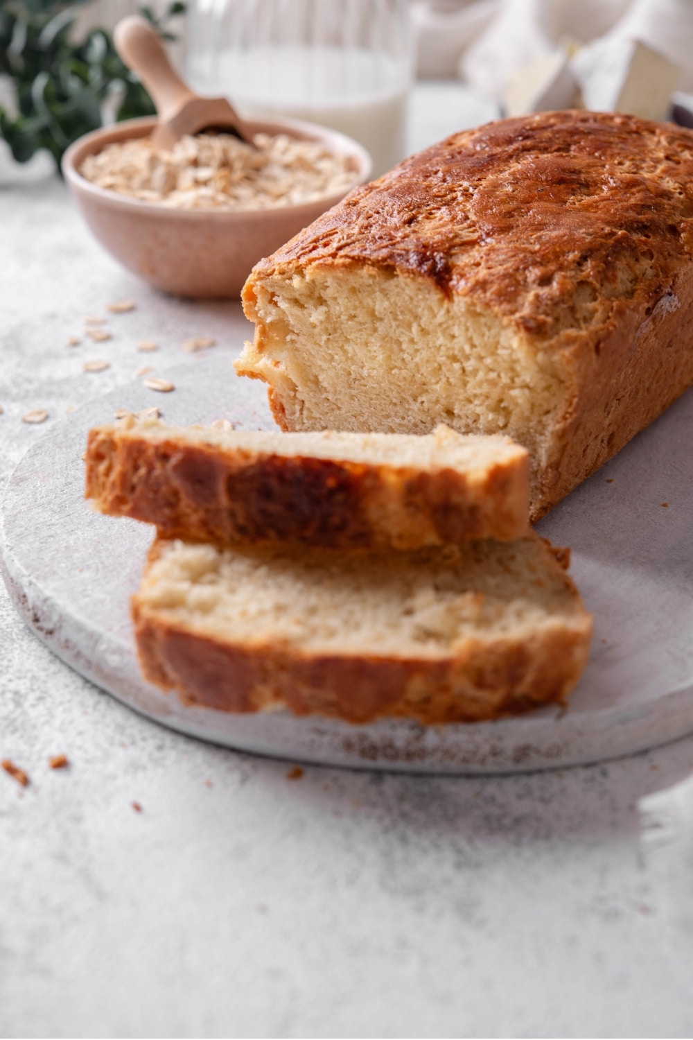 One loaf with two slices of oat bread sit on a wooden server. In the background there is a bowl of oats with a scooper inside it.