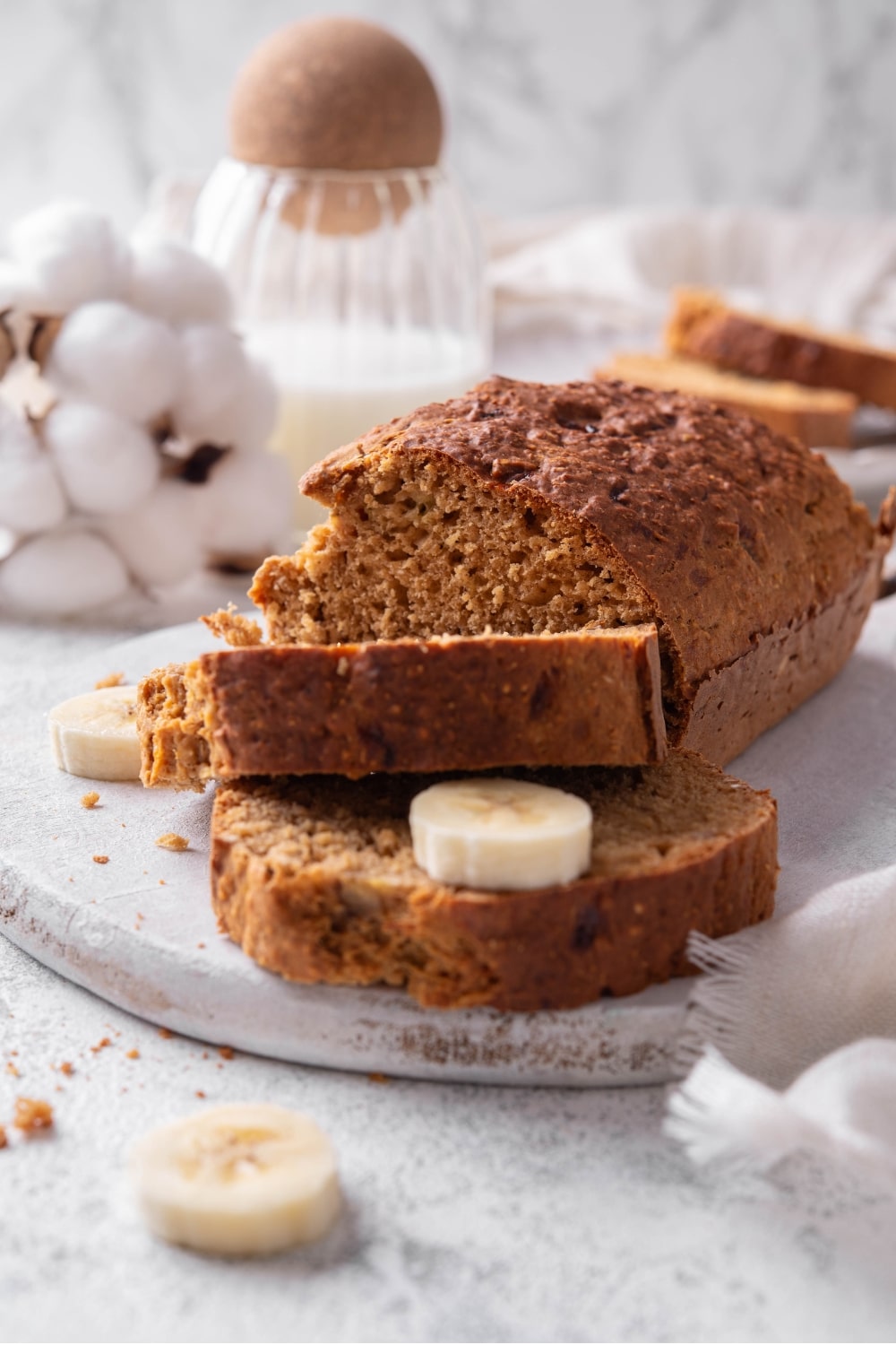 A low calorie banana bread loaf and two slices of banana bread sit on a white serving board. There is a piece of banana in between the two slices, and additional pieces of banana on the board.